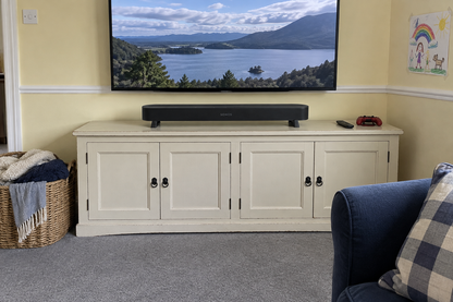 A white wooden media console cabinet with four doors in a living room setting, supporting a black soundbar and a large wall-mounted TV showing a lake landscape. A blue armchair is partially visible on the right, and a wicker basket with blankets is on the left.